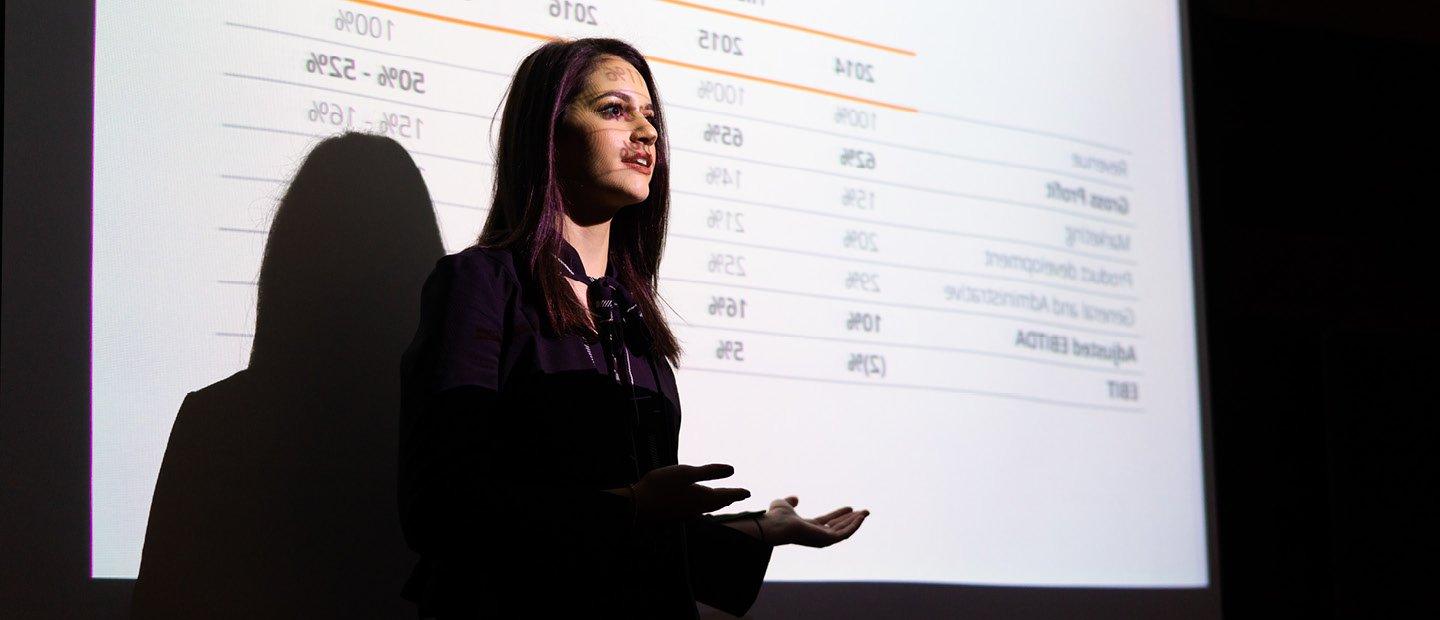 A young woman standing in front of a projector screen displaying percentage data.