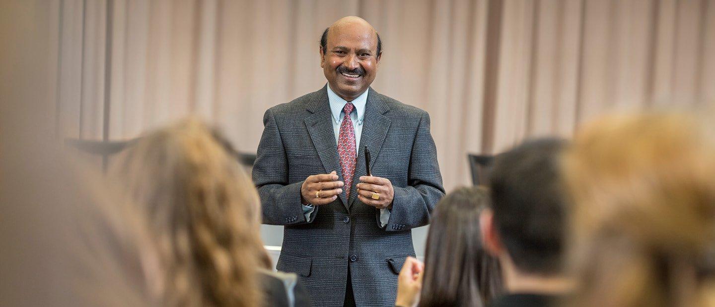 A professor in a suit, facing the camera, smiling at a room full of students.