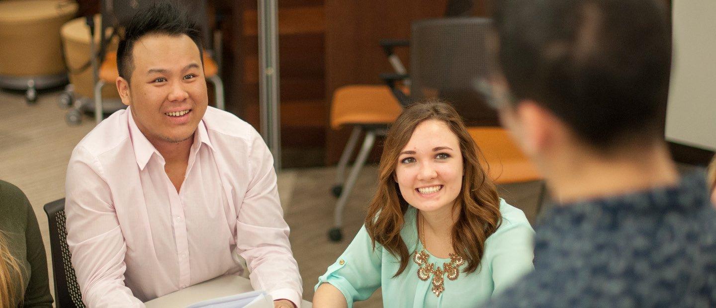 A man and woman seated at a desk, looking up and smiling at a man standing in front of them.