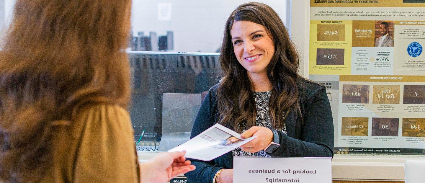 A woman standing in front of a graphic poster of statistics, handing a flyer to another person.